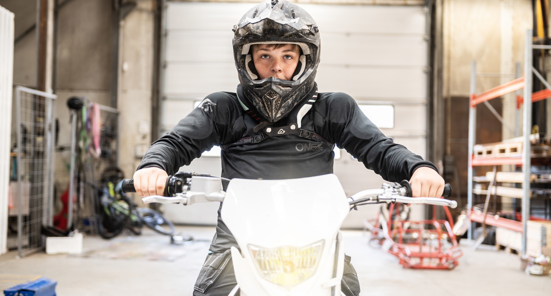 Elev i motocrosshjelm sidder på hvid motorcykel i værksted. 