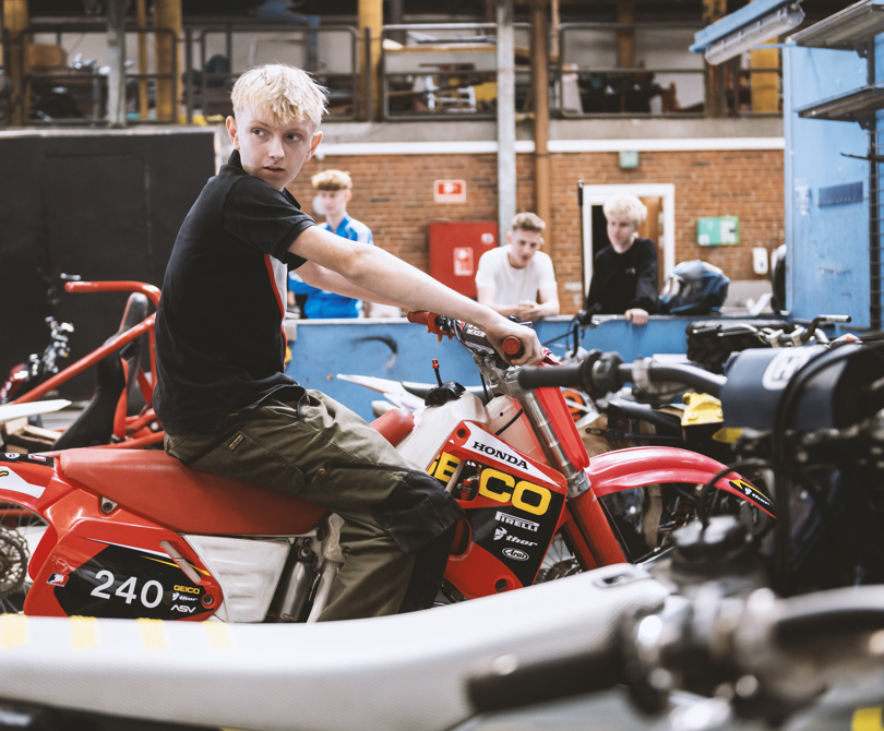Elev sidder på motocrossmotorcykel i skolens værksted. 