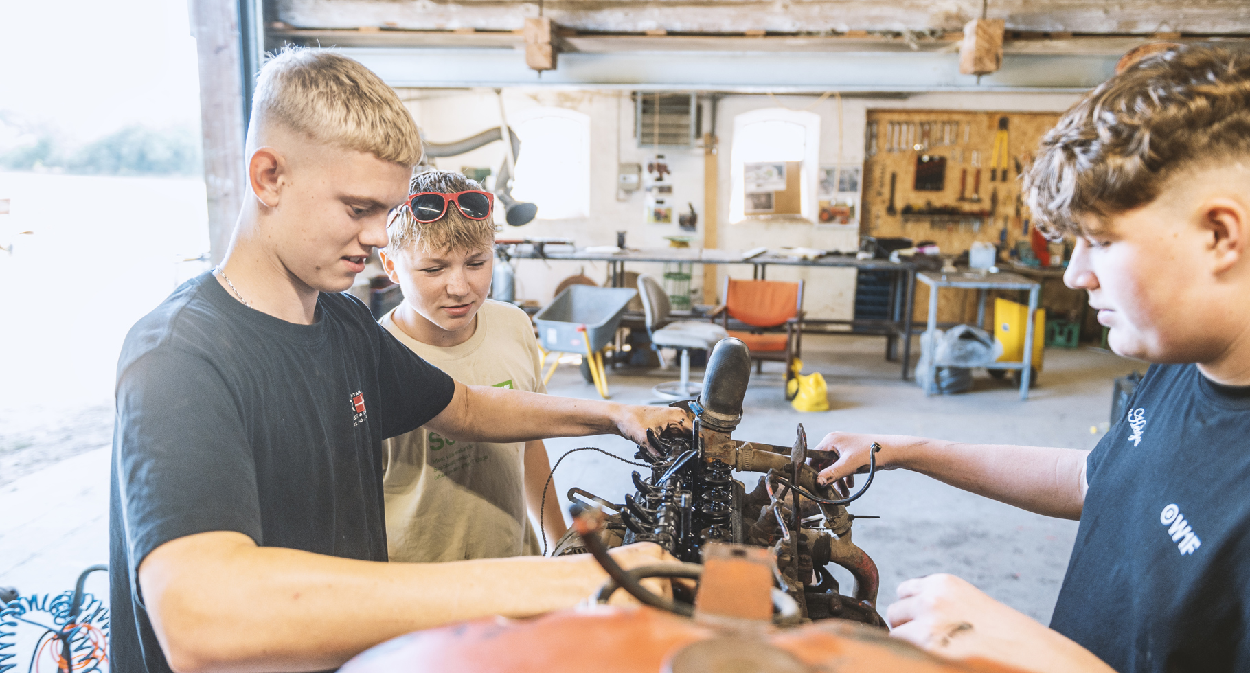 Tre elever arbejder sammen på traktormotor i værksted.