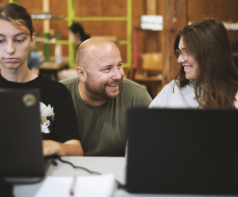 Lærer smiler til elever ved computere i klasseværelse.